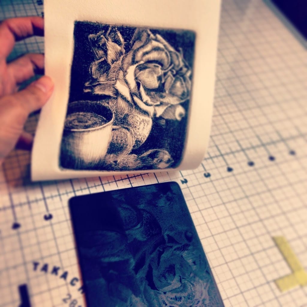 pulling a drypoint print from a sheet of black plexiglass after a trip through a printing press, where the line work on the plate has transferred to the paper revealing a still life of a rose, a cup and a bird figurine